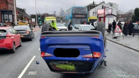 A blue Skoda Fabia resting on its head after crashing. The car is in the middle of a congested road. There is a bus in the foreground.