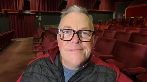 Richard Knights/BBC Wayne Burns smiles at the camera while sitting inside the cinema on red chairs. He has short grey hair and wears black glasses with a black and red puffer jacket. 