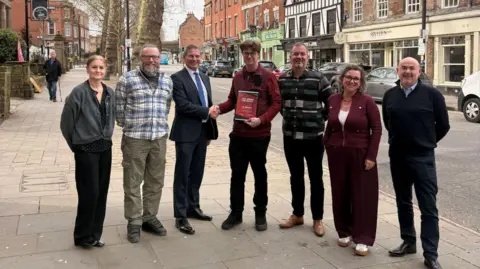 Supplied A group of people stood in a row on a pavement. Jon is in the middle holding his award and shaking hands with a man in a suit.