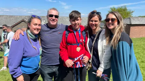 A family group of five people dressed in outdoor clothes. Ben is pictured in the middle wearing his medal over a red hoodie and holding a Union flag
