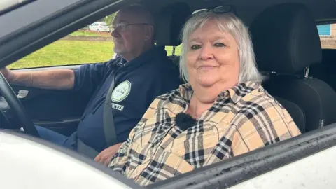 Evie McLean sitting in the passenger seat with volunteer Danny Prew with his hands on the steering wheel. Evie, wearing a checked beige shirt is smiling. 