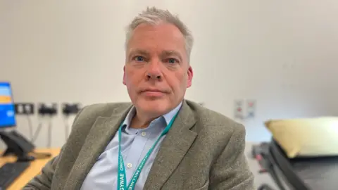 Somerset NHS Foundation Trust A close-up of David Milliken in a clinical setting wearing a smart jacket, blue open-necked shirt and turquoise lanyard. There is a computer and a patient bed behind him