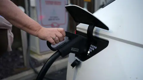 A close up of a person's hand holding a charger for an electric vehicle and putting it into the socket of a white car. In soft focus in the background, a charging point is visible.