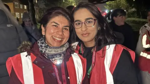 Two women looking into the camera and smiling wearing high-vis pink jackets. The one on the left smiles, showing her teeth and has long dark hair, tucked behind her ears. The other has dark rimmed glasses on and smiles but with her lips closed.