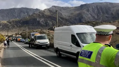 The back of a police officer (right) in high vis uniform and hat standing on the A5 in the area of Llyn Ogwen, several vans line the road, including tow trucks taking cars away. The mountains of Eryri cut through the sky in the background. 