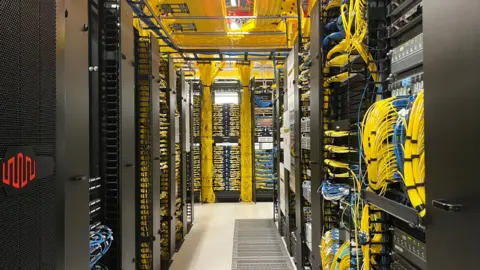 A corridor in a warehouse. To the right, lots of yellow cables are running in and out of various machines. To the left, there are some large black panels and more wiring. 