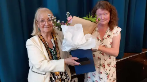 Councillor Sue Eacock, left, pictured with Councillor Joanna Kane when she became the new Mayor of Redditch - Councillor Eacock is wearing a white jacket with a large gold chain around her neck. She is being handed flowers to mark the occasion.