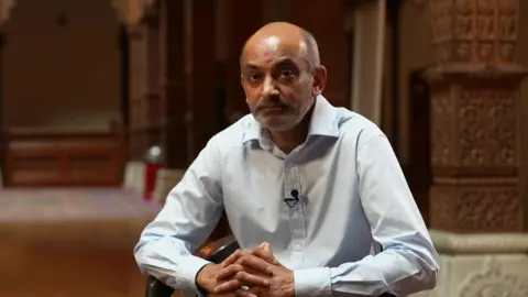 Tarun Patel is seen a light blue shirt sits inside a Hindu temple, hands clasped, with carved pillars and soft lighting in the background.