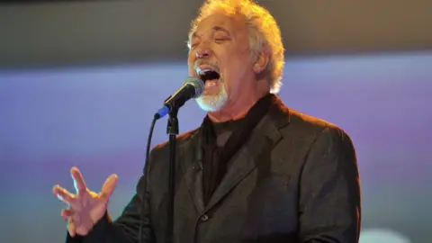 Singer Tom Jones, mid song, singing into a mic, gesticulating with eyes closed