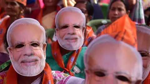 Women wearing Narendra Modi masks, and saffron stoles of the Bharatiya Janata Party, and some wave BJP flags, at an election campaign in Assam's Nagaon district on 25 March 2026.