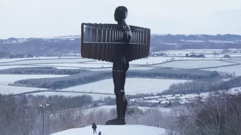 PA Media People sledging in the snow at the foot of Antony Gormley's Angel of the North sculpture in Gateshead. The tall sculpture, which resembles a man with rectangular wings, is dusted with snow.