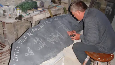 John Candler John is sitting on a stool in his workshop with a very long piece of grey stone laid out in front of him, on which he has carved some cartoon figures and the phrase, 'too little told responsible for so much'. There is a very dusty bench behind the stone with lots of tools and other articles