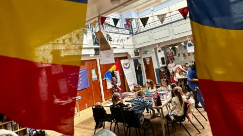 The blue, yellow and red of the Romanian flag can be seen in the foreground with children sitting around a desk in the background. 