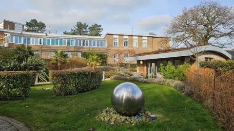 St Luke's Hospice A modern building in the distance overlooks a green lawn and shrubbery. There is a wooden building to the left and a large stainless steel sphere in the middle of the grass.