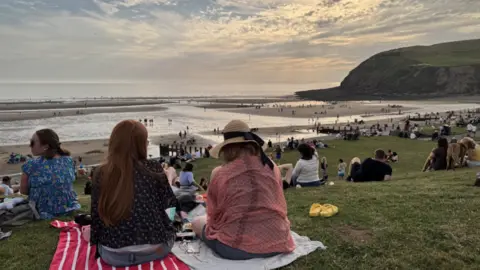 Ed Hanson/BBC The sun sets on St Bees Beach, where groups of people are gathered en masse across the grassy dunes or the wet beach itself.