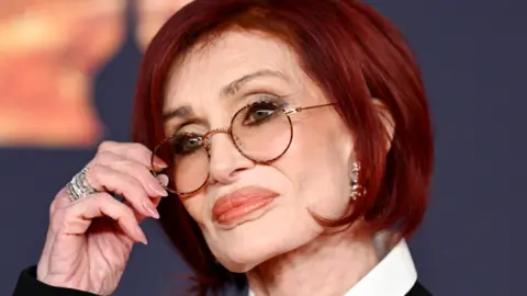 Getty Images A woman with bright red hair and one hand holding her wire framed glasses