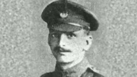 Grainy black and white photo shows a young man with a neat moustache wearing a military cap and uniform.