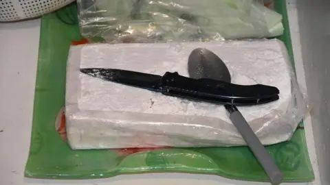 Cleveland Police A knife with a black handle sits on top of a dessert spoon. Both utensils are sitting on top of a black of white powder wrapped up in a plastic sheet on a kitchen counter top.