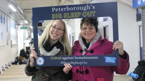 Light Project Peterborough Davina wearing a pink coat, standing with a colleague. They are smiling at the camera through a cardboard Big Sleep Out sign that has the middle cut out.