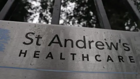 Dawid Wojtowicz Silver sign with black letters which say 'St Andrew's Healthcare' in black letters. It is located outdoors and rainwater can be seen on its surface.