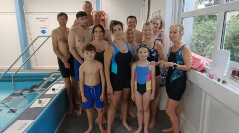 Laura Alliston The group of volunteers, all ranging in age and gender. The youngest is six and the oldest is 69. They wear swimming trunks or costumes and stand determined at the edge of the pool.