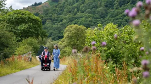A man and a woman make their way along a section of the Keswick to Threlkeld Trail. The main is on a mobility scooter and is wearing a navy cap, navy body warmer and navy trousers. The woman to his left is walking. She has grey hair and glasses and is wearing a blue dress. Trees, shrubs and flowers are well established on either side of the path.