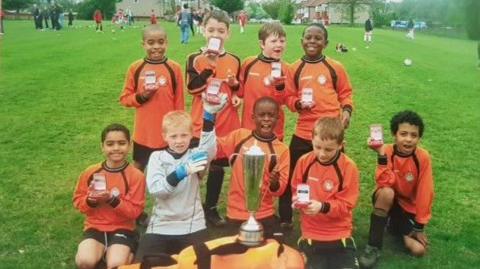 A youth team at Cray Wanderers celebrate a trophy, with a young Marc Guehi holding the trophy in the centre
