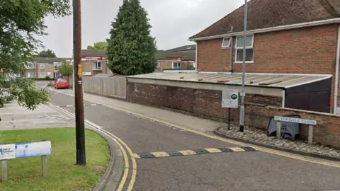 Silverdale Close, which has yellow and black speed humps and small blocks of flats .There is a tall conifer tree and a red car in the distance. 