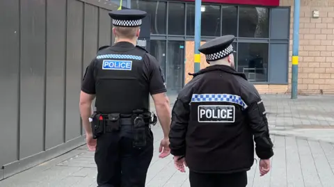 Two police officers on patrol in a pedestrianised area. They have their backs to the camera and are wearing their police caps and uniforms.