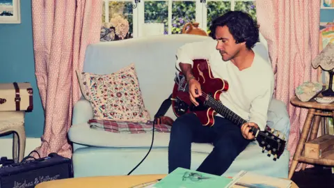 Chris Floyd Jack Savoretti sitting on a blue couch in a living room, playing an electric guitar. There is a lot of furniture around him, such as tables, vases and cases.