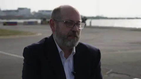 Nick Hancock is pictured on a tarmacked waterfront site in Southampton. He is bald with glasses and a full beard and wears a dark jacket over a white collared shirt.