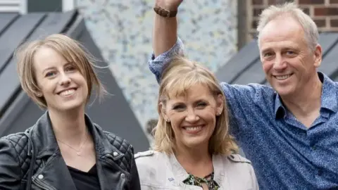 PA Media Laura Nuttall smiles as she stands next to her parents. She is wearing a black leather jacket and a black top and she has blonde hair in a bob.