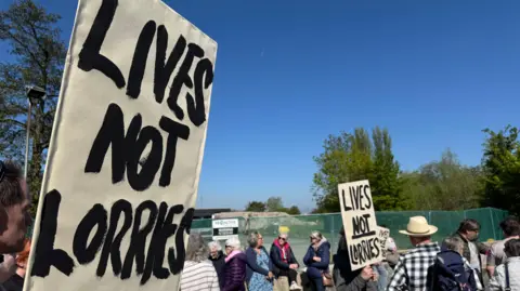 A campaigner holds a sign that says "lives not lorries" in front of the site of the proposed warehouse. In the background, more campaigners are gathered. One of them holds a sign with the same text.