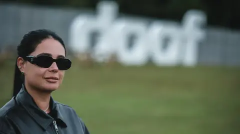 Graeme Ogston Hannah Laing, in dark sunglasses, her dark hair in a ponytail and wearing a black leather jacket, stands with a large "Doof" sign behind her in Camperdown Park