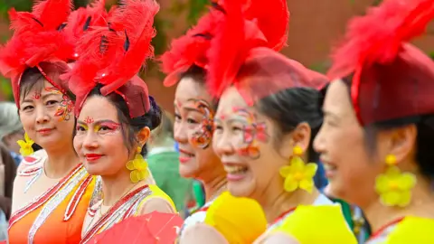 Pacemaker Five women are wearing bright yellow and orange clothing. They have red hats on with feathers. 