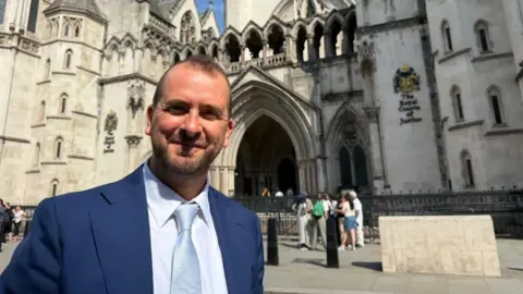 A man is standing outside a court building. He looks smart, wearing a blue jacket and light blue tie. 
