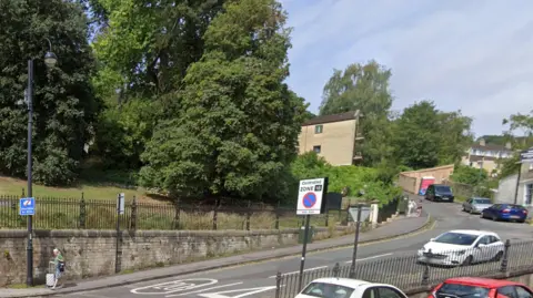 An image of a street leading up a hill, a park is visible in the back ground. Cars are parked by the curb and iron railings separate the pavement from a drop on to the street below, which is a few feet lower down.  