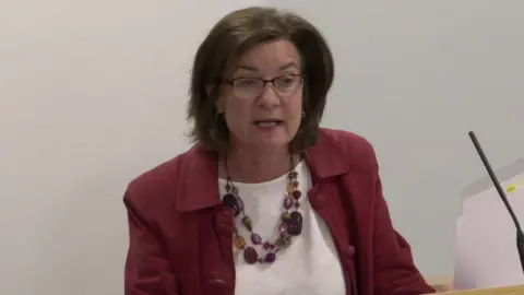 Eluned Morgan stood in the Senedd's temporary debating chamber wearing a red jacket, white top, a colourful necklace and glasses.