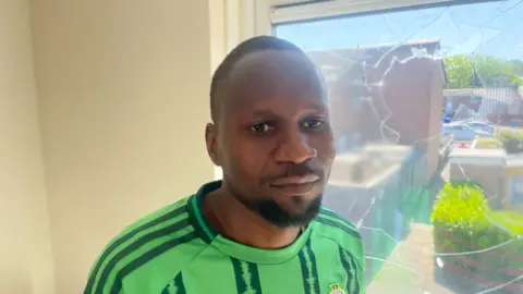  Mutasim Mohammed. He has short black hair and beard. He is standing in front of a smashed window on his home. He is wearing a Northern Ireland football top