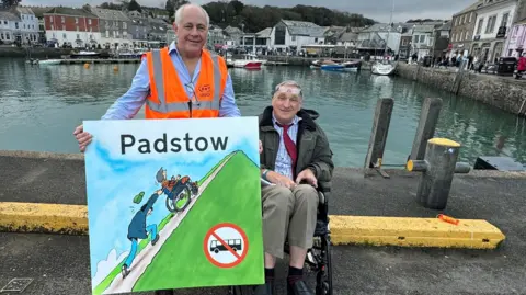 RMT Barry West stands with David Redgwell who sits in a wheelchair beside Padstow harbour. Mr West holds a large sign that shows an illustration of a man struggling to push a person in a wheelchair up a steep hill and there is a 'no buses' sign.
