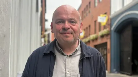 A bald man with grey stubble, he is smiling towards camera, wearing a grey shirt and navy jacket. The background is out of focus, red bricked buildings in Belfast's Cathedral Quarter