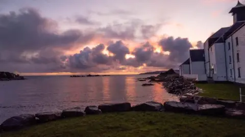 Paul Gorman Una escena costera al atardecer con un edificio blanco a la derecha, una costa rocosa y nubes coloridas sobre aguas tranquilas.