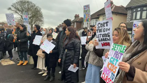 A group of men, women and children all wearing coats are stood at the side of the road holding placards