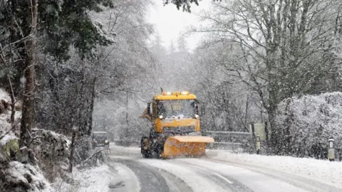 PA Media A bright yellow snow plough driving along a road in a blizzard ploughing the road 