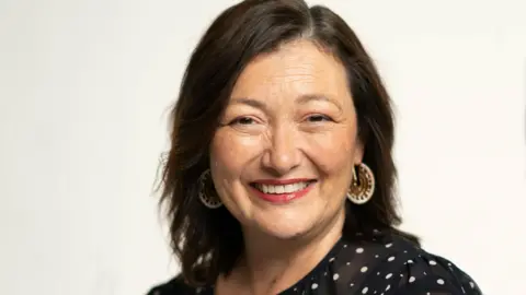 Birmingham Royal Ballet/ Johan Persson Caroline Miller pictured smiling with shoulder length brown hair, brown eyes and red lipstick. She is wearing gold hoop earrings and a black sheer top with white spots on it.