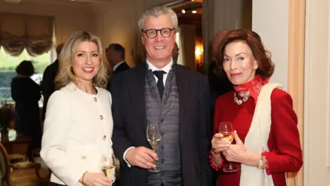 Getty Images A man standing between two women, all three are well dressed and smiling at the camera while holding glasses of white wine.