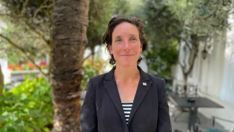 Deputy Lindsay de Sausmarez - A woman with short brown hair and blue eyes looking at the camera. She has a black blazer on with a black and white striped top underneath. She is wearing a Guernsey pin badge on her left lapel.