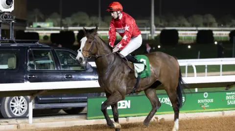 Lo Chun Kit/Getty Images Oisín Orr riding Spirit Dancer on a racecourse in Saudi Arabia, in 2024. Or is wearing red and white silks with a star pattern on the sleeves, a red helmet and white Jodhpurs. The chestnut horse has a white stripe on his face and is wearing a green racing saddle. A black 4x4 vehicle is parked at the side of the race track. 