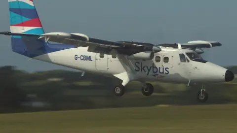 A white Skybus plane with a blue and red fin tail heads in to land at an airport.
