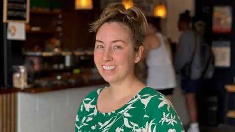 Heather is smiling at the camera. She has her hair tied up and is wearing a green and white dress. She is in the cafe in Northstowe. There are out of focus customers in the background, buying coffee from the barrista.